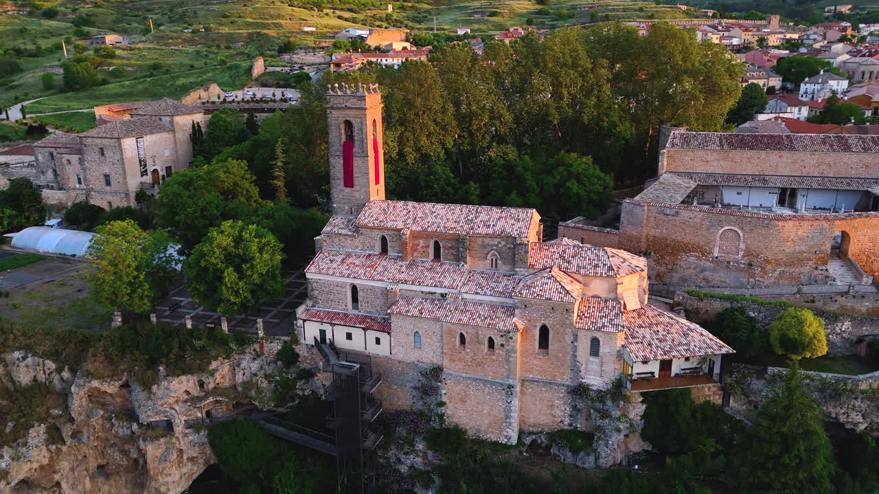 Drone orbit around the Romanesque church of Brihuega, La Alcarria, Spain, perched on a cliff. The clip shows historic stonework, bell tower, cemetery walls and golden sunrise light over the village
