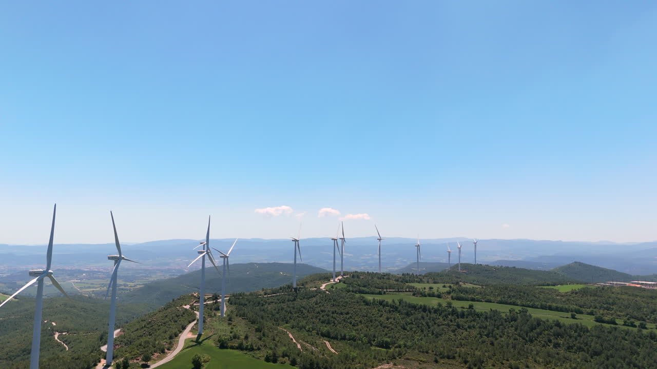 Aerial clip flying parallel to a row of wind turbines spinning on forested hills. Clear sky, clean energy and scenic mountain landscape captured by drone