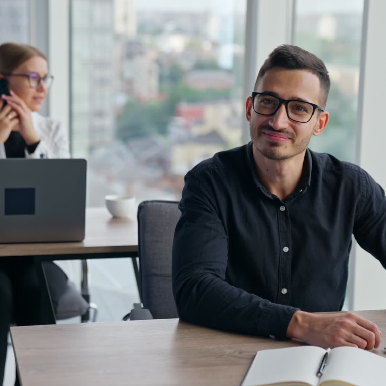Positive smiling male specialist working in the office. Portrait of an average office employee working at laptop. Female speaking on the phone at backdrop