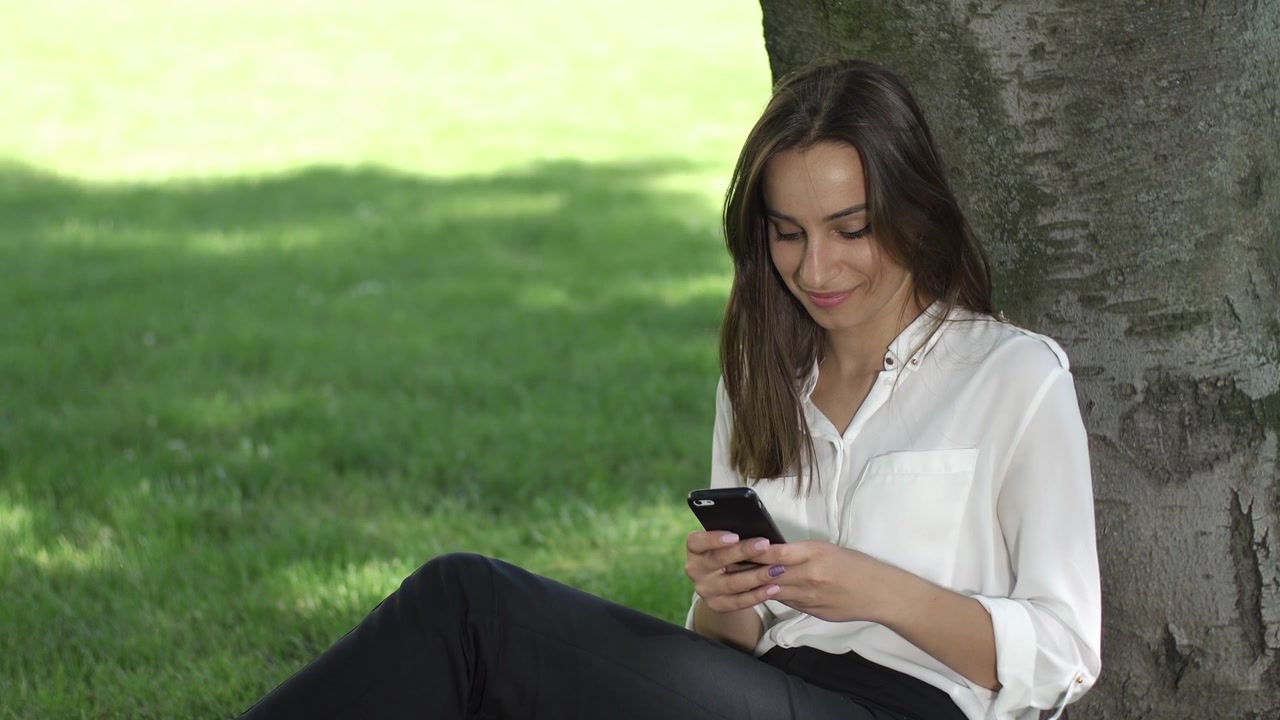 Giovane donna si rilassa sotto un albero in un parco, usando lo smartphone.