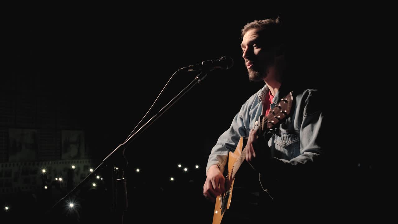 한 뮤지션이 큰 무대에서 기타를 연주하고 있으며, 관객들은 배경에 불을 들고 있습니다. 관객실은 아름다운 박이는 불빛으로 가득 차 있습니다.