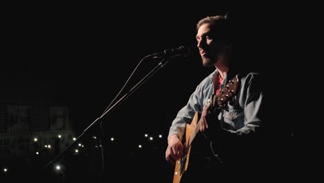 a musician is playing guitar on a large stage with audience members holding flashlights in the background. the auditorium is filled with beautiful flashing lights.