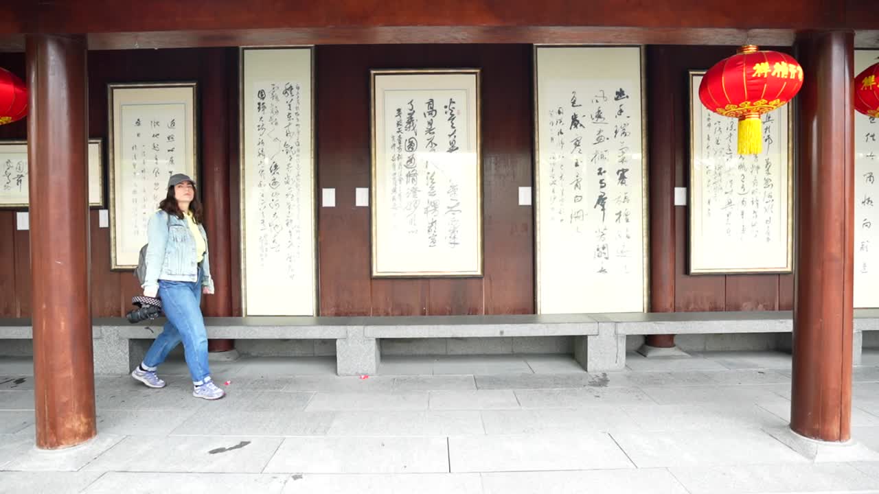 Caucasian tourist woman with a camera walks along a corridor with Chinese proverbs and red lanterns at Longhua Temple, Shanghai, China