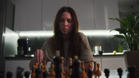 Close-up-a-serious-brunette-girl-in-a-beige-T-shirt-plays-chess-for-one-of-the-pieces-at-a-black-dining-table-near-a-home-plant-in-a-modern-kitchen-in-the-evening