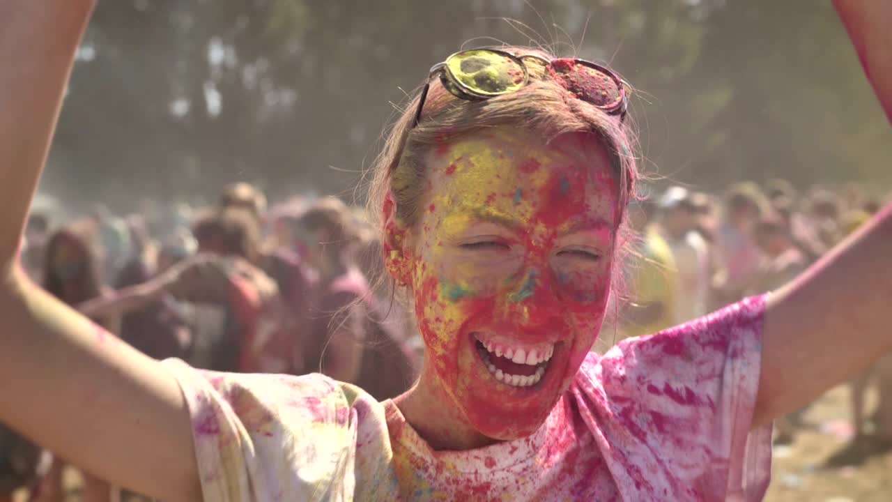 Young happy girl in colourful powder is histerically laughing and dancing on holi festival in daytime in summer, watching at camera, color concept, emotional concept