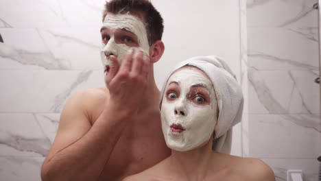 pareja loca y divertida de pie frente al espejo en el baño después de la ducha - hombre topless, chica con toalla blanca en la cabeza - haciendo caras divertidas, posando, haciendo muecas con máscara blanca en la cara