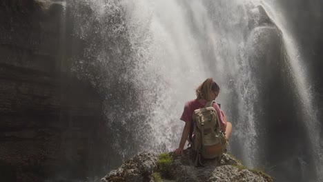 Una-Chica-De-Excursión-Está-Sentada-En-Una-Roca-Bajo-Una-Cascada-En-Una-Reserva-Natural