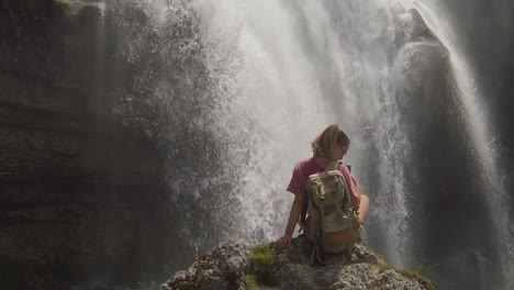una chica de excursión está sentada en una roca bajo una cascada en una reserva natural