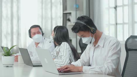 a man of three asian call center agents wearing headset and mask speaking to customers on the call while two of his colleagues are talking during working at the office