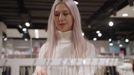 cheerful woman with pastel hair and lavender manicure in chunky knit sweater strolls to brightly lit rack, gently brushes multicolor polos on hangers, inspecting fabric and hue