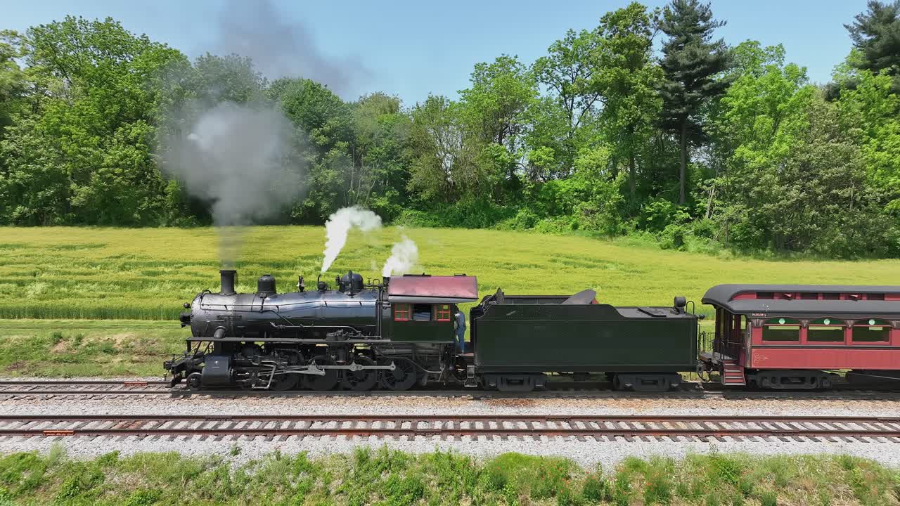 A vintage steam train travels slowly through lush green countryside emitting smoke into the clear sky smooth motion rich detail vibrant colors cinematic quality