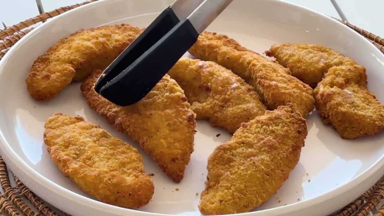A hand uses black kitchen tongs to pick up and move golden-brown breaded chicken tenders on a white plate in bright, natural lighting
