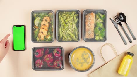 Food delivery top view, take away meals in disposable containers. Lunch boxes with cooked vegetarian dishes, using phone with chroma green screen. Healthy diet. Catering service