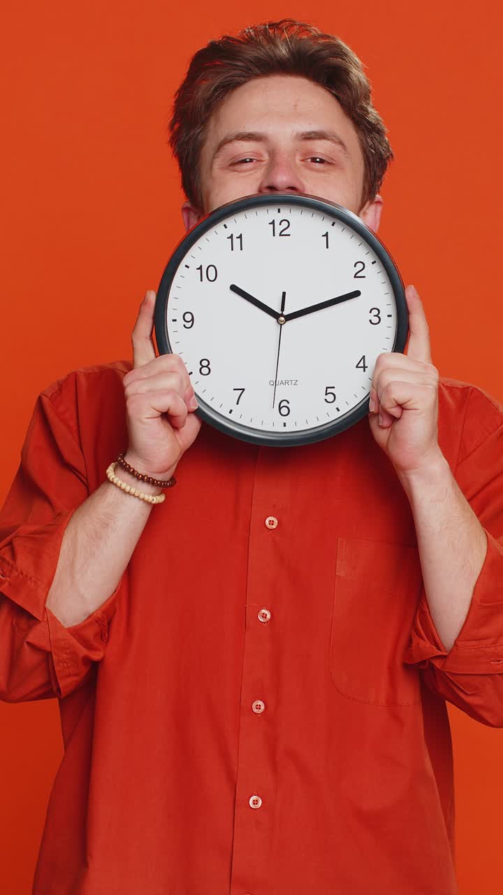 Serious strict young man holding office clock hiding behind checking time on watch obscuring face