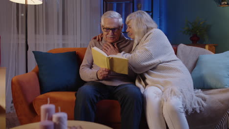 senior grandparents couple relaxing, reading book, talking enjoying leisure hobbies at night home