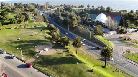Aerial-view-of-the-landscape-in-the-park-with-a-road-while-cars,-the-theater-and-the-sea-pass-in-the-background-on-a-sunny-day-in-Montevideo,-Uruguay