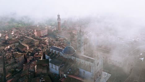 Aerial-over-the-Duomo-di-Siena-in-Siena,-Province-of-Siena,-Italy-in-early-morning-fog