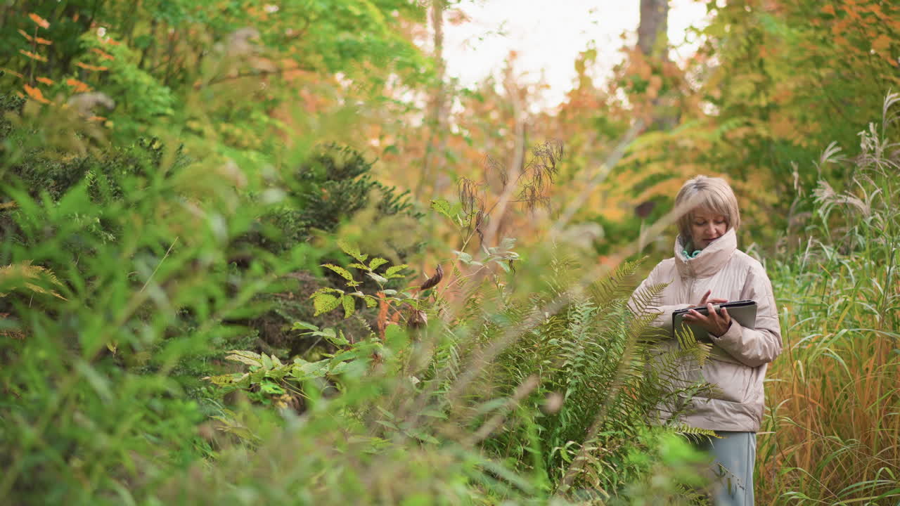 botanist holding tablet inspects vibrant green fern leaves in dense woodland area with golden foliage, documenting plant characteristics during serene moment of autumn field research