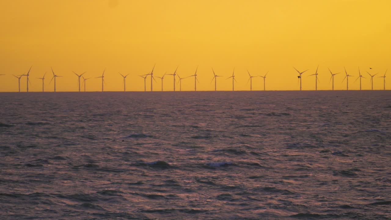 Beautiful Orange Sunset Lighting Silhouetting Offshore Wind Turbines Farm Generating Renewable Clean Green Electricity in United Kingdom. Modern Technology Creating Energy. Sea Waves in Foregorund