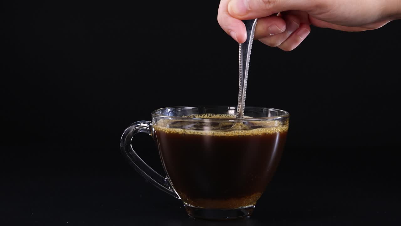 A hand stirs sugar into a glass cup of tea against a black backdrop, highlighting the mixing process