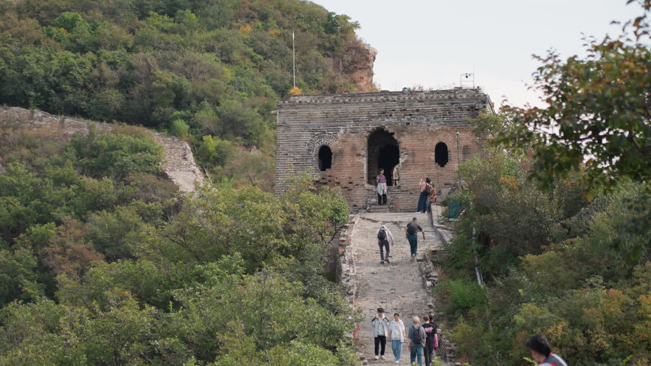 Cancello in pietra su una collina con visitatori che salgono un sentiero attraverso una fitta vegetazione, antico arco in mattoni incorniciato da alberi, gruppo di escursionisti che salgono gradini irregolari verso una fortezza in rovina sotto un cielo pallido, calma