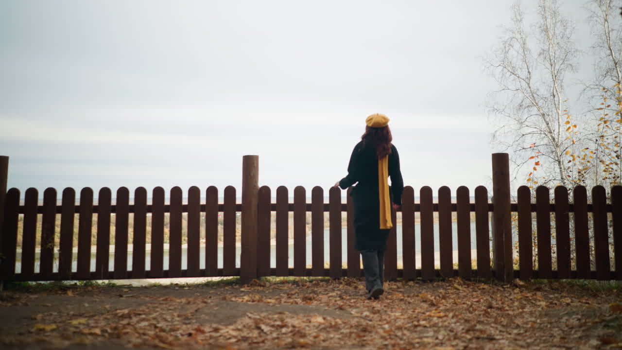 노란색 베레트와 검은색 코트를 입은 여자가 나무 울타리를 향해 걸어가며, 그 위에 손을 고, 건조한 가을 잎이 어져있는 울타리를 통해 강의 전망을 감상합니다.