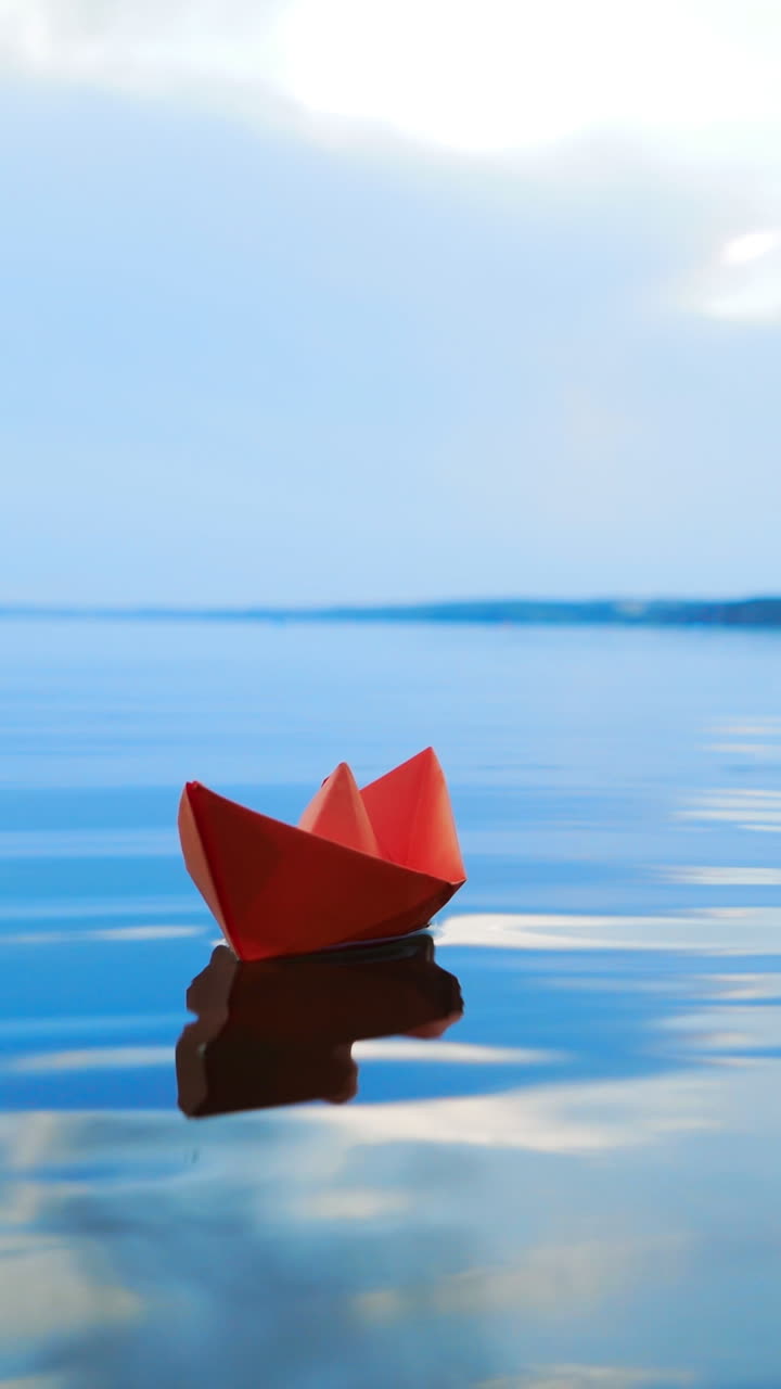 Ship of red paper floating in the blue river at clear day under the blue sky. One paper boat swims on the calm lake in summer Vertical video