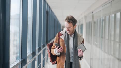 Happy-adult-man-walking-down-a-glass-corridor-receives-a-joyful-notification-on-a-smartphone