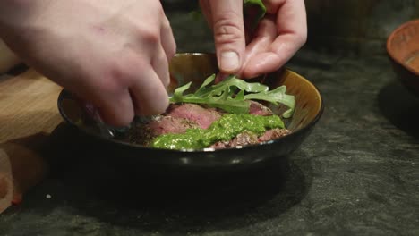 chef plating bbq meat dish with green vegetable grass in dark ceramic bowl