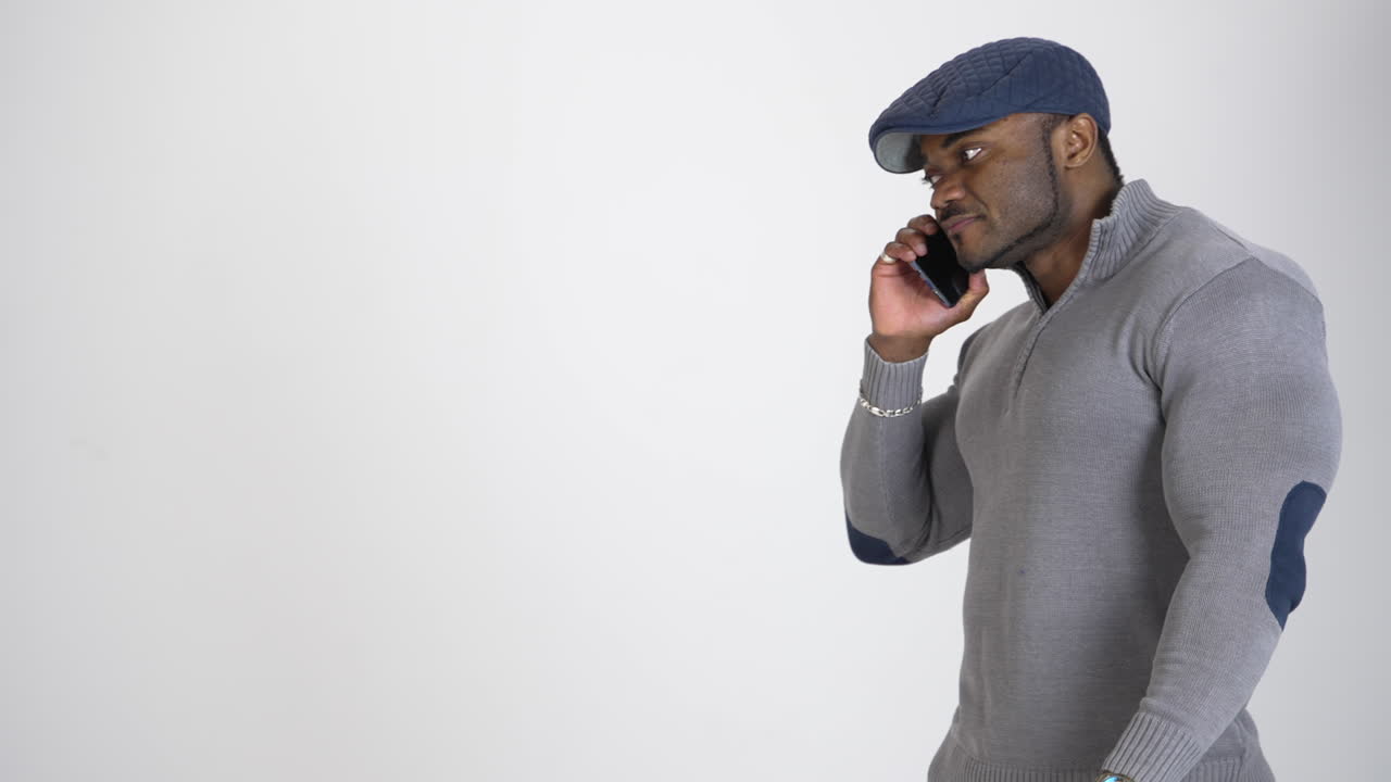 Young african american man speaks on phone. Black guy in blue cap and grey sweater talking smart phone on white studio background. Profile view.