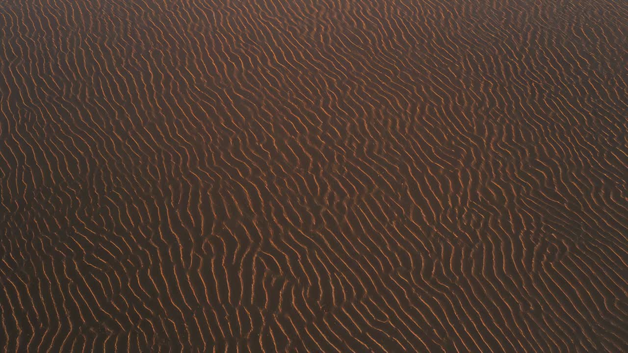 Descending drone capturing closer view of rippled sand dunes at sunrise, highlighting sunlit ridges