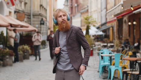 a young man with a beard wearing headphones walks down a busy street in a european city.