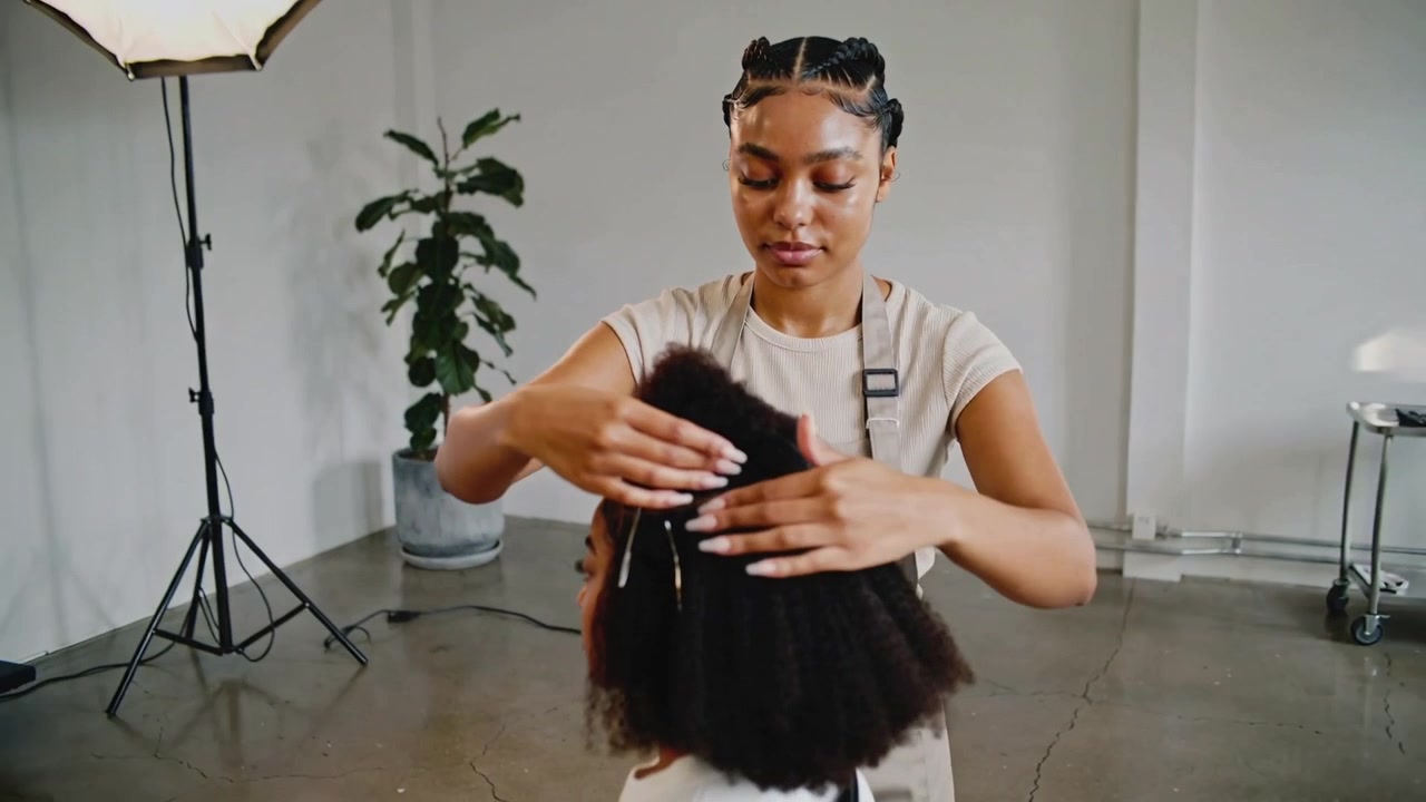Holding top section stylist forming bun on child-client's hair at studio wearing apron and clips