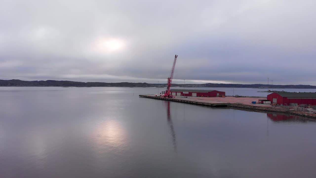 Flying towards the red construction warehouses of Lysekil by the calm, peaceful waters of Sweden on a cloudy day - Aerial shot