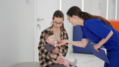 Over-the-shoulder-of-a-confident-brunette-girl-a-pediatrician-in-a-blue-uniform-a-doctor-playing-with-the-little-daughter-of-a-brunette-girl-in-a-checkered-shirt-in-a-modern-clinic