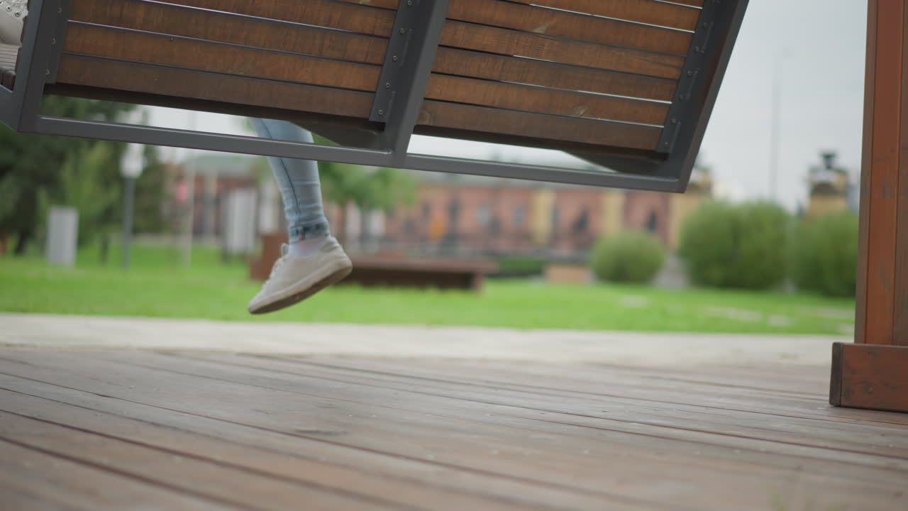 Jeans og sneakers, der dingler under bænken på trædækket i en park. Nærbillede i lav engel, der fanger fodens bevægelse og afslappede tempo, en ensom, legende stemning, overskyet vejr og planker med tekstur