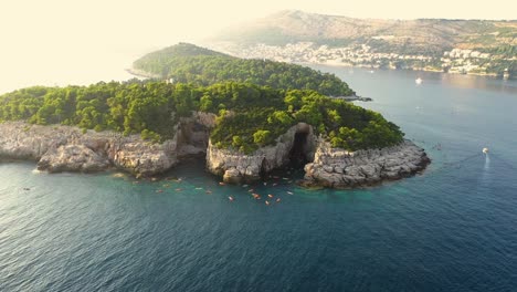 Aerial-footage-of-the-Lokrum-Island-and-a-group-of-tourists-on-a-kayak-tour-and-a-boat-passing-by,-Dubrovnik-,-Croatia