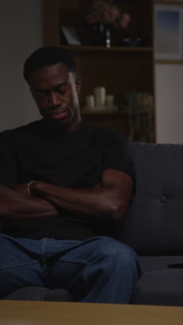 vertical video of unhappy and depressed young man sitting on sofa at home looking anxious and worried resting head on hand 1