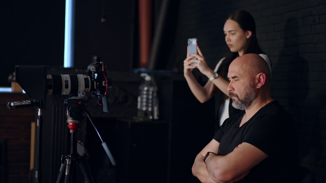 People working backstage in photo studio. Focused bearded cameraman sits behind his camera looking at its display. Woman takes video on her phone.
