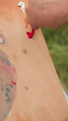 un primer plano de un momento de colaboración en la creación artística, con la mano de una mujer sosteniendo una paleta de madera mientras la mano de un hombre exprime cuidadosamente la pintura roja sobre ella