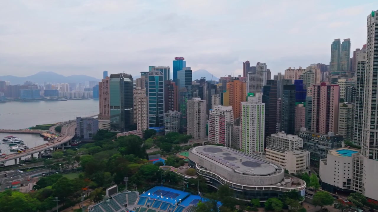 Luchtopname met terugtrekkende pan onthult Victoria Park zwembad, tennisstadion en blauwe banen richting de dichte hoogbouwappartementen en wolkenkrabbers van Causeway Bay met de haven van Hong Kong daarachter.