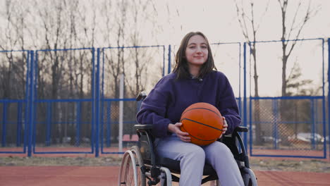 Glückliche-Behinderte-Frau-Im-Rollstuhl,-Die-In-Die-Kamera-Blickt,-Während-Sie-Einen-Basketball-Auf-Dem-Basketballplatz-1-Hält