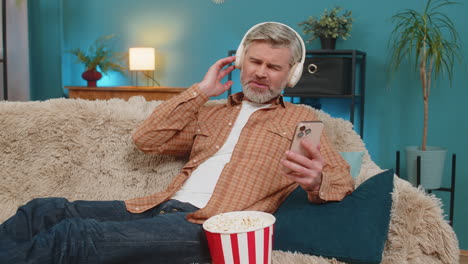 mature man sitting on sofa with phone wireless headphones eating popcorn listening to music at home