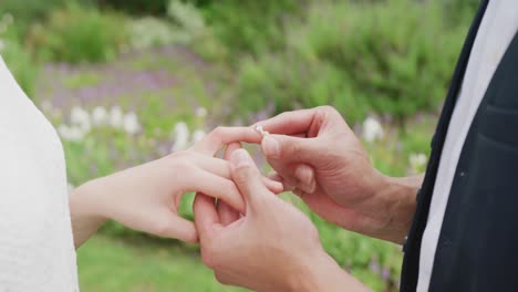 Diverse-couple-holding-hands-and-putting-ring-on-sunny-day-at-wedding