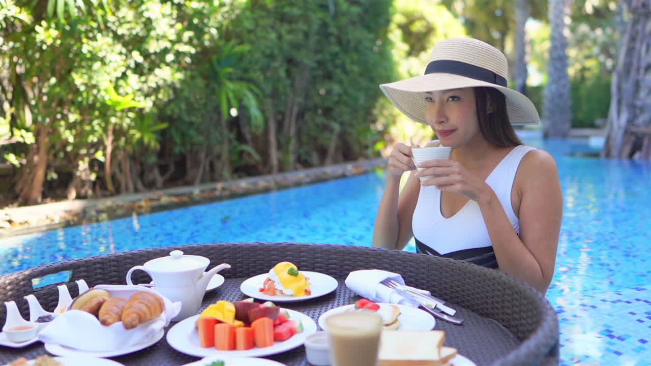 A young Asian woman in a pool chooses coffee from a floating platter of breakfast foods