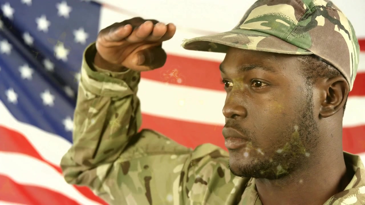 Soldato in uniforme mimetica che batte le palpebre, alzando la mano destra per il saluto militare davanti a uno sfondo con la bandiera americana.