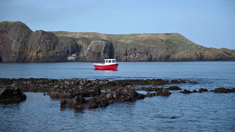 Static-wide-shot-of-red-Fishing-boat-cruising-on-ocean-water-with-gigantic-mountain-range-in-background---Blue-sky-and-sunlight-during-daytime