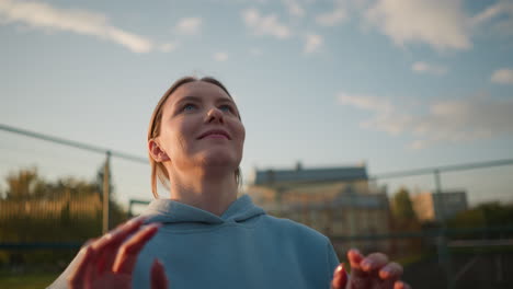 close-up of lady in blue sweater playing volleyball outdoors, arms raised with a joyful expression, greenery and buildings create a scenic background under a bright sky