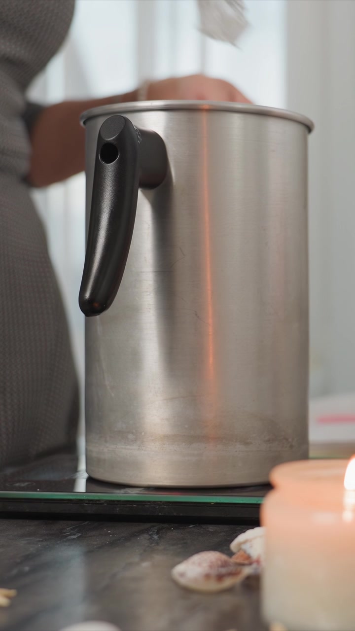 Nahaufnahme eines Edelstahlbehälters mit schwarzem Griff auf einem Induktionskochfeld. Eine brennende Kerze und dekorative Muscheln sind im Vordergrund sichtbar, was eine ruhige Atmosphäre suggeriert. Die Szene ist weich fokussiert.