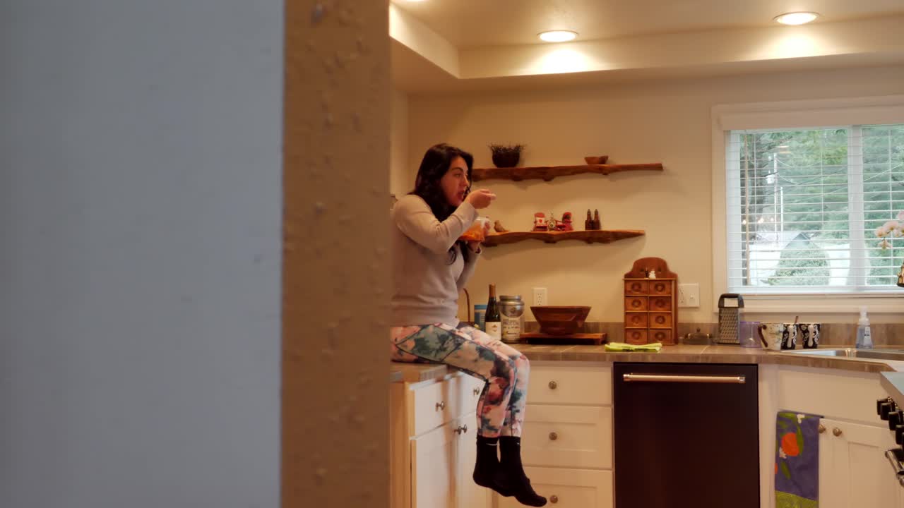 mujer hispana comiendo sobras del recipiente tupperware, sentada en la encimera de la cocina